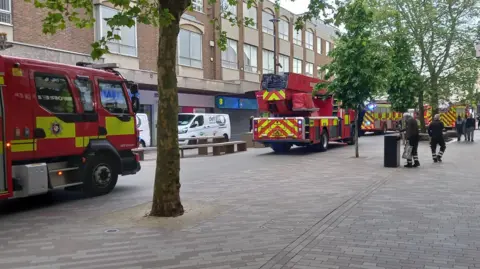 Supplied Four fire engines parked in the middle of a town centre between trees and shops. Pedestrians are walking alongside them. 