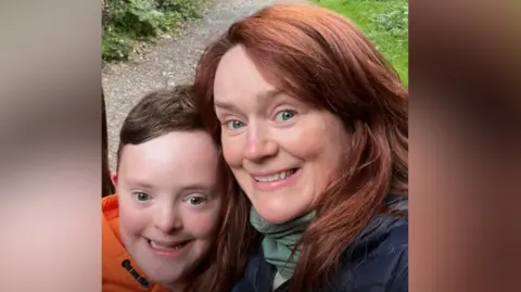 Family handout Finn has brown hair and wearing an orange hoodie. Angela has red hair and wearing a navy jacket with a green scarf. They are standing on a path surrounded by trees and smiling at the camera.