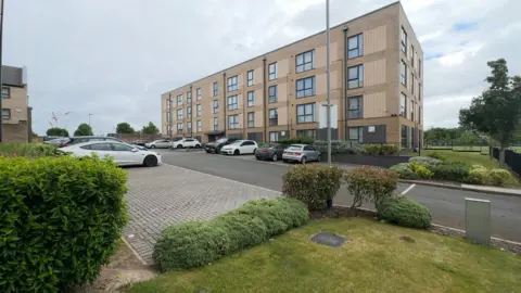 Wide view of a modern four-storey brick apartment building with large rectangular windows, set beside a paved parking area with several parked cars. In the foreground, there are neatly trimmed shrubs, low hedges, and a small grassy area. A streetlamp stands near the building, and additional residential structures and trees are visible in the background under an overcast sky.