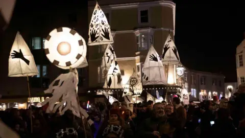 Getty Images A parade of smaller lanterns held up by a crowd making its way through the streets. They are passing the buildings that line the streets in the night time scene.