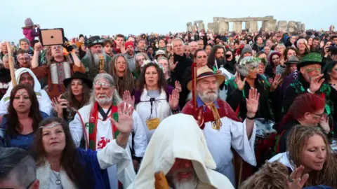 PA Media Druids and pagans wearing traditional clothing are among a crowd of thousands with Stonehenge in the background against a grey sky.