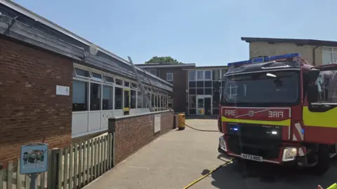 A fire engine is parked outside the school building. There is a ladder leaning up against the side of the building leading to the roof. A hose from the engine is also leading on to the roof.