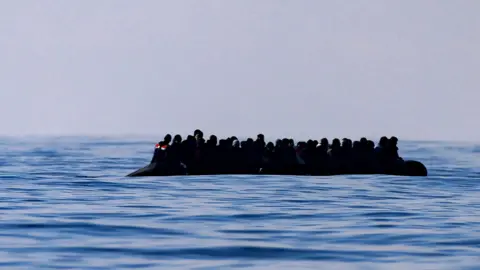 A inflatable dinghy carrying around 65 migrants can be seen silhouetted against a grey sky on a blue sea, as it crosses the English Channel in March