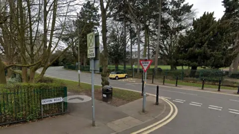 A junction next to a park wit tall trees. A give way and zone ends road sign are on the corner.  