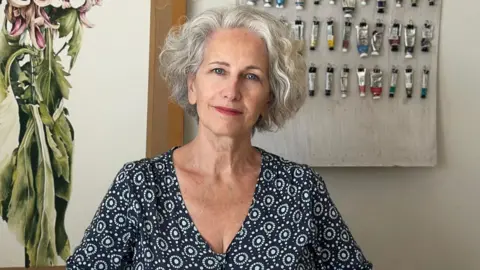 Marie Burke is sitting in front of a wall with paints hanging from nails. She is wearing a patterned shirt and has grey hair.