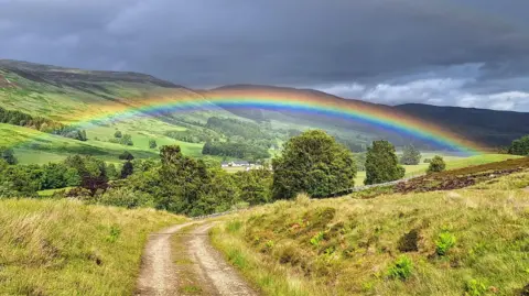 Bruno Delatousche A rainbow with a number of colours over a dirt track surrounded by grass verges leading towards a cluster of trees. Beyond the trees is at least one building, which is brightly-coloured. The sky is a mixture of dark grey colours.