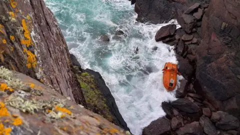 An upside down orange lifeboat wedged in some dark rocks with white water all around it