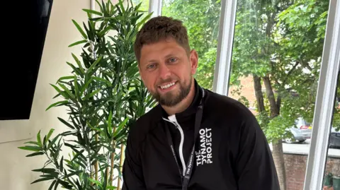 A man with short brown hair. He is smiling and wearing a black hoodie that reads "The Dynamo Project". He is sitting in front of a window and next to an indoor green plant. 