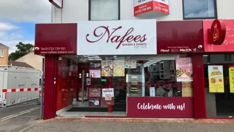 JOANNA TAYLOR/BBC Maroon shop sign that reads 'Nafees Bakers and Sweets' above glass-fronted shop. There are posters on the glass and a sign which says 'Celebrate with us!'. To the left of the shop there is a silver gate fencing off a road.