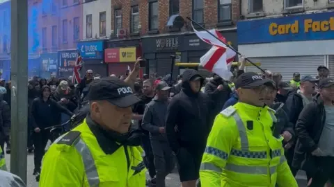 A large group of protesters, flanked by police officers in reflective yellow jackets, march along a street lined with shops, all of which have their shutters pulled down. Some protesters are carrying English and British flags, and some blue smoke from a flare is visible hanging in the air to the left.