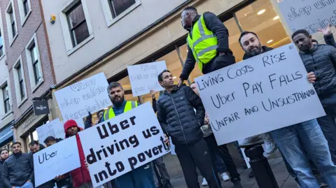 A group of Uber drivers holding signs saying "Uber driving us into poverty" and "Living costs rise, Uber pay falls, unfair and unsustainable".