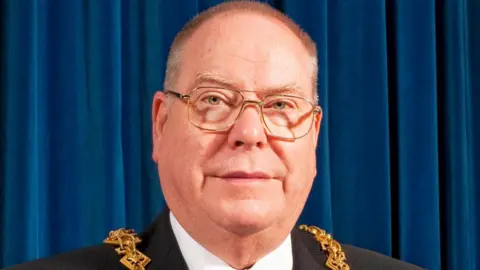 A man in his 60s with short brown hair and glasses wearing a suit with a ceremonial chain