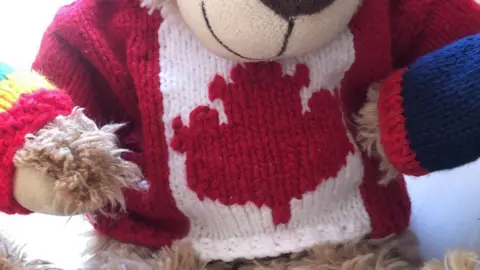 Stuart Race A close up of a small knitted shirt being worn by a teddy bear. It represents the Canadian flag with stripes of red and white along with a red Canadian maple leaf.