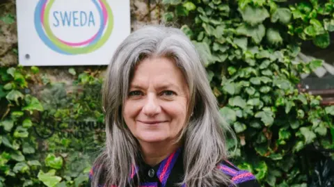 Alice Florrie Paula wearing a pink and black top, with long grey hair standing in front of an ivy covered wall and a white sign with her charity's logo on it.