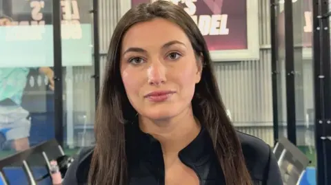 A woman is wearing a black jacket. She has long brown hair. Behind her are two Padel courts, and a framed sign. 
