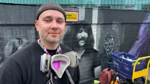 Mr Murals next to his murals in Birmingham. He is in front of the work in progress of a a 120ft (36.5m) mural depicting all four original members of Black Sabbath. He has a purple mask hanging around his neck. There is also a trolley with paint cans nearby. 
