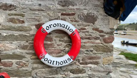 A red and white lifebelt with the words 'Coronation Boathouse' attached to a stone wall with the river to the right and a boat moored in it.