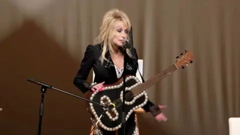 Dolly Parton sings into a microphone with a bedazzled guitar hanging from a strap astir   her neck