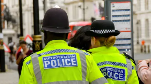 Two Metropolitan Police officers patrol a street in hi-vis jackets. Their backs are turned towards the camera. 