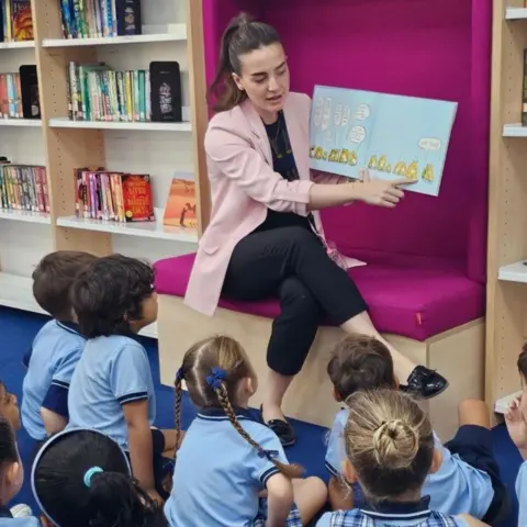 Louise Fraser Louise Fraser is reading to young children in a classroom. She is sitting facing them and has a book open so they can see it. There are shelves with books on behind her. The children are wearing light blue T-shirts. 