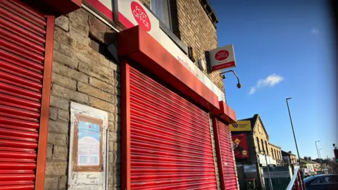 A post office on a busy high street. The red shutters are all down and there is a closure notice visibly posted on the wall.