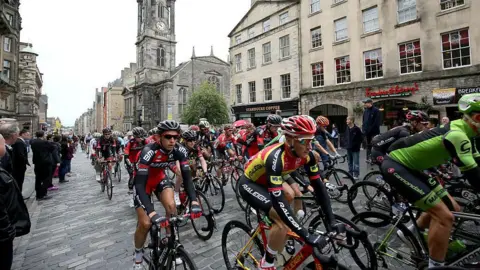 A group of cyclists, known as a peleton, travelling from left to right on the Royal Mile in Edinburgh. There are people watching by the side of the road. A Starbucks and the Tron Church can be seen in the background.