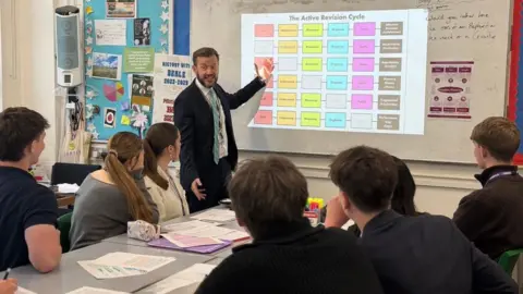 Simon Beale A man wearing a suit pointing at a whiteboard with revision information to a group of students wearing black blazers. 