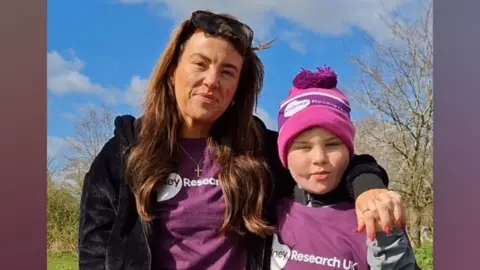 Sammy, a woman with shoulder length dark brown hair, with Caelan, a boy wearing a purple T-shirt and woolly hat. They are on their walk at Ferry Meadows in Peterborough.