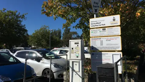 A parking machine with a sign next to it that says Garras Wharf Long Stay Car Park. There are hatchback cars parked in the car park. 