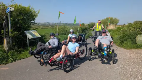 Four people on recumbent trikes smile for the camera. They are out on the Exe Estuary Trail on a sunny day.