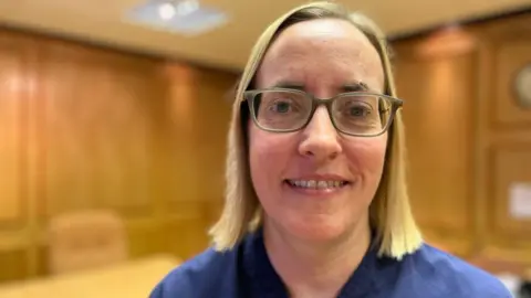 Helen has shoulder-length blonde, straight hair. She is wearing glasses and a navy blue NHS uniform.