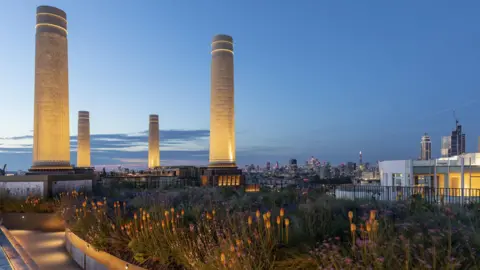 A shot at night of Battersea roof gardens, with flowers lit up and the power stations famous chimneys in the background. 