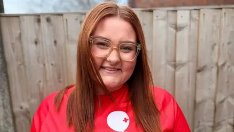 Libby Reynolds Libby stands in front of a garden fence with her bright red London Marathon t shirt on.