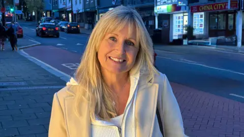 A woman with shoulder length blond hair wearing a white zip too and cream overcoat smiling with a road and shops behind her. 