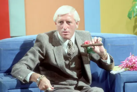 Jimmy Savile smoking a cigar and wearing a grey suit, sitting on a blue sofa