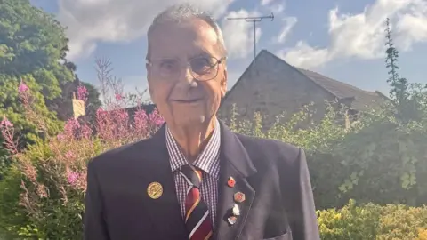 Supplied A man in a navy suit, gingham shirt and striped tie. There are Remembrance poppy badges on the lapel of his jacket. He is wearing glasses and has short grey hair. He is standing outside with plans and flowers behind him, and a house can be partially seen behind the plants.