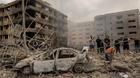 Joel Gunter/BBC A car is coated completely in grey dust and surrounded by rubble, while a group of men survey the scene over damaged buildings.