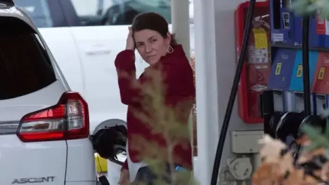 Mega Agency Anne Sacoolas with long dark hair tied back. She has her left hand touching her forehead and her right holding a petrol pump with which she is filling a white Subaru Ascent car. She is wearing a dark red jacket. There is another white car behind her.
