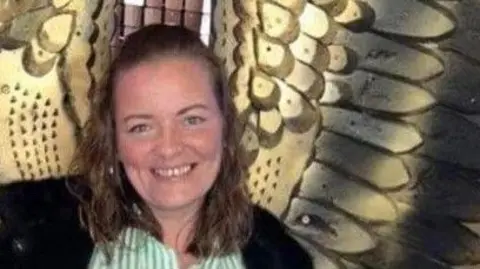 Dianne Cleary pictured in front of a wall with a large bronze set of angel wings on it. She is smiling and wearing a stripey green and white shirt and open black coat. Her wavy hair is half pinned back from her face.