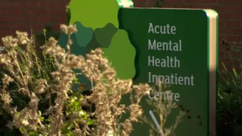 Signo de la BBC que decía "Centro impaciente de salud mental aguda" Detrás de algunos arbustos frente a una pared de ladrillo rojo.