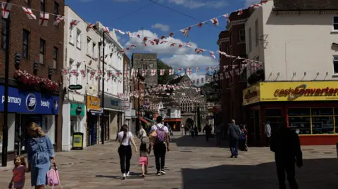 Dartford High Street. There are shops including Boots, Specsavers and Cash Converters lining the pedestrianised street which has bunting of English flags above.