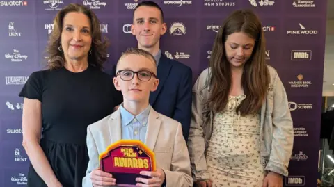 Bernie Davis Four people are stood in front of an advertising board. On the left is a woman with brown hair and wearing a black dress. To her left his a young boy in a pale suit wearing glasses and holding an orange Into Film awards trophy. Behind him is another boy in a dark suit and with dark short hair. To his left is a young girl wearing a flowery dress and long brown hair, she is looking down and not at the camera like the others.