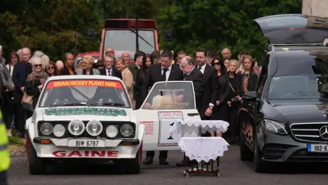 PA Media A white rally car with the name GAINE emblazoned on the bottom of it sits in front of a crowd of mourners in black. Behind a tractor can be seen.