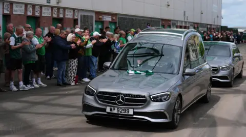 A silver hearse with the coffin of Sir Tom Farmer drives past supporters gathered outside Easter Road Stadium in Edinburgh