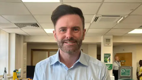 A man with short dark hair and a trimmed beard stands facing the camera in a bright office environment, wearing a light blue button-up shirt. The background shows a drop ceiling with vents and lights, desks and equipment, wall posters, and a doorway where another person and signage are visible.