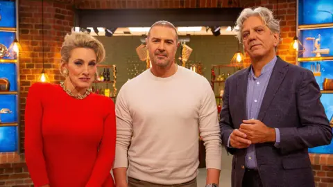 A woman with blonde hair, styled up, wearing a red jumper, a chunky gold necklace and large earrings, stands next to a man with short hair and facial hair and a cream jumper, with another man with grey hair, a blue shirt and a dark blue blazer to their right. They are stood in front of a kitchen set with brick walls and various cooking equipment and ingredients on shelves.