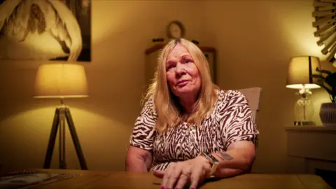 Yeti Television/BBC Cymru Wales Cathy Broomfield is a woman in late middle age. She is white and has dyed blonde chest length hair. She is wearing an above the elbow sleeved dress with a brown and cream patterned dress. She is seated behind a table with her left hand resting on it. She wears an large square watch and a wedding ring. She has a tattoo on her lower left arm. She is sitting in a living room with a mantlepiece to her right with a lamp on it, and a standard lamp to her right. There is an out of focus cabinet behind her and a painting on the wall.