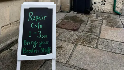 A chalk A-board sign outside a stone building that says 'Repair cafe 1-3pm, bring your broken stuff'