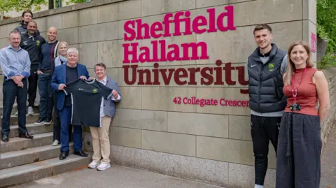 Six men and three women are standing either side of a large Sheffield Hallam University sign which is red and pink lettering on a stone wall.  Seven are standing to the left-hand side, up a set of steps. Two are holding a black football shirt with a grey neckline