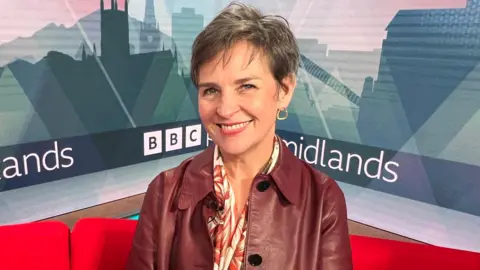 A woman wearing a red leather jacket and a floral scarf smiles at the camera. She is sitting on a red sofa with 'BBC Midlands' signage in the background. 